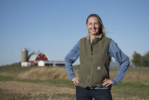 A young woman stands, smiling, in a farm field with hands on hips.